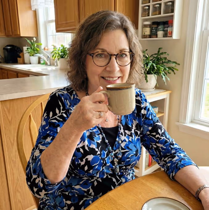Mujer sonriente con lentes y blusa floral azul, sentada en una mesa de cocina de madera con una taza beige.