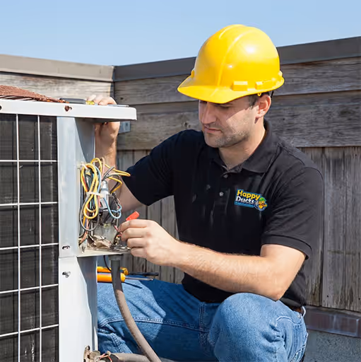 Técnico con casco amarillo y camisa negra reparando una unidad HVAC exterior con cables expuestos.